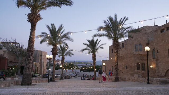An old city view with modern lights, vehicles, trees and galleries at dusk. here, there's a wide stairs with people walking. the distant beach is also visible. May 23rd, 2022, Jaffa, Israel.
