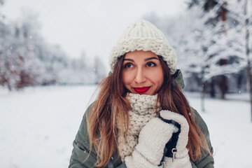 Fototapeta na wymiar Close up portrait of happy young woman in snowy winter park wearing warm knitted clothes and red festive lipstick.