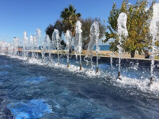 Fresh water fountain with jets in a pool with a beach background and trees on a sunny day.
