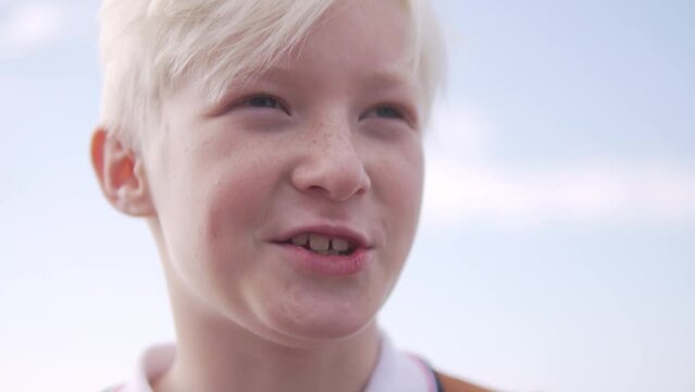 A blond boy looks at the camera against the sky and talks.