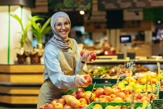 A young beautiful hijabi Muslim woman, a supermarket worker, lays fruit in the department. He holds apples in his hands, looks at the camera, smiles.