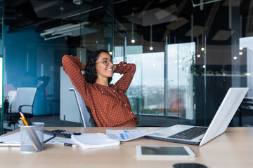 A break at work. Successful work result. A young beautiful Latin American woman sits smiling in the office at the desk, hands behind her head, resting on a chair.