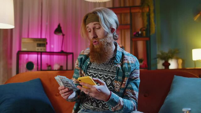 Planning family budget. Bearded redhead man counting money dollar cash use smartphone calculate domestic bills at home. Joyful hippie guy satisfied of income saves money for planned vacation, gifts