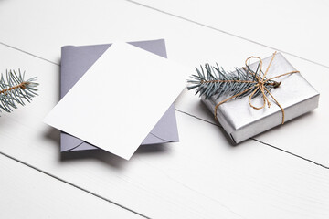 Christmas greeting card mockup with grey envelope, silver color gift box and green fir tree branch on white wooden table. Empty winter holiday card
