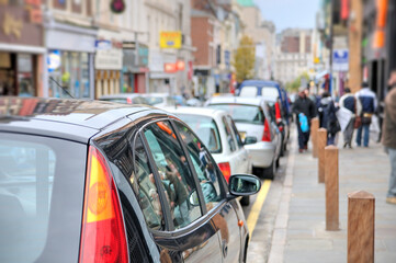 A typical busy high street scene in the UK with parked cars and pedestrians.