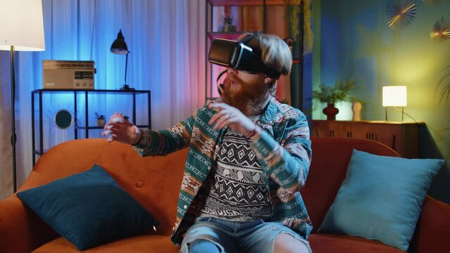 Bearded redhead man using virtual reality futuristic technology VR app headset helmet to play simulation 3D 360 video game, watching film movie at modern home apartment. Guy in goggles sitting on sofa