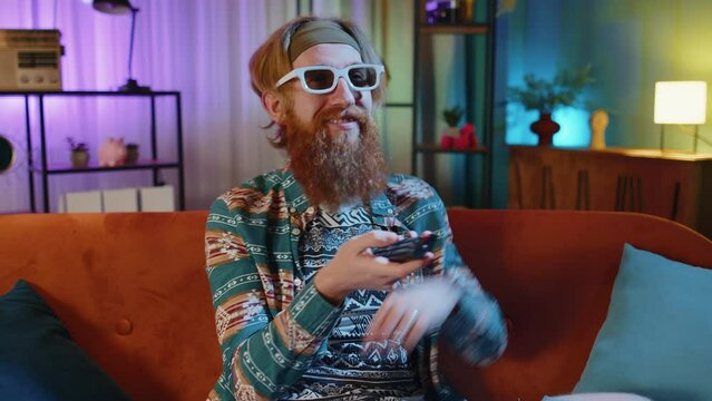 Excited bearded hippie young man sits on sofa use TV remote control watching serial, sport game, film, online social media movie content at home. Guy enjoying domestic entertainment, eating snacks