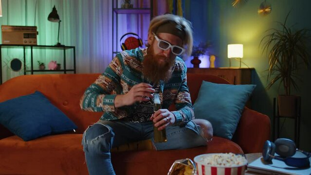 Happy bearded hippie young man sits on sofa drinking beer eating chips snacks and watching TV serial, sport game, film, online social media movie content at home. Guy enjoying domestic entertainment