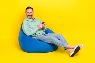 Full length photo of positive cheerful guy dressed green hoodie sitting bean bag chatting instagram twitter telegram isolated yellow color background