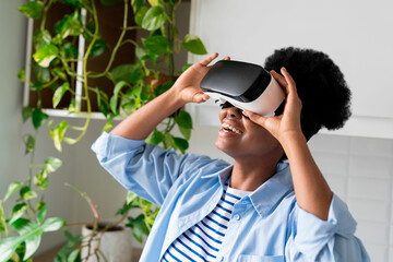 Inspired African American woman with VR goggles on eyes looking up standing in room with closet. Smiling Brazilian girl suffers from video game addiction and loves to spend time in virtual metaverses