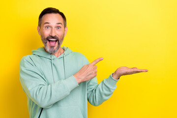 Photo of mature middle age excited man grey stubble finger point hold arm empty space new brand sweatshirt isolated on yellow color background