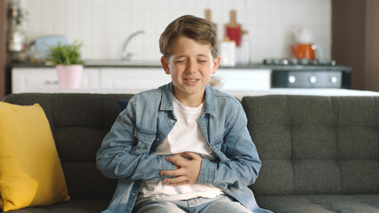 Little boy suffering from stomachache while sitting on sofa at home.The child has a stomach ache. - obrazy, fototapety, plakaty