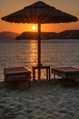 View of a sun bed, a sun umbrella and a stunning orange sunset at the beach of Mylopotas in Ios Greece