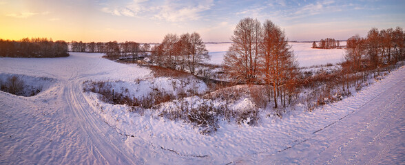 Winter panorama. Snow covered fields, meadows. River, frozen trees, village in evening light. Rural sunset landscape. Dirt road in ice