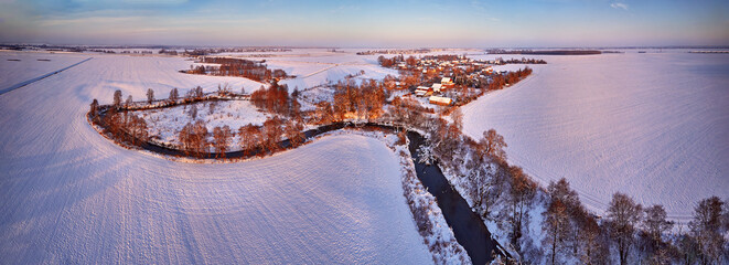 Winter panorama. Snow covered fields, meadows. River, frozen trees, village in evening light. Rural sunset landscape. Dirt road in ice
