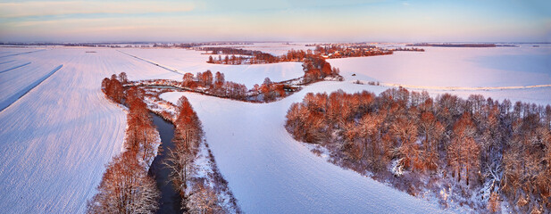 Winter panorama. Snow covered fields, meadows. River, frozen trees, village in evening light. Rural sunset landscape. Dirt road in ice. Aerial view