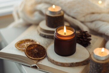 Cozy autumn or winter composition with aromatic candle, wool sweater, fairy lights, book. Aromatherapy, home atmosphere of cosiness and relax. Wooden background close up.