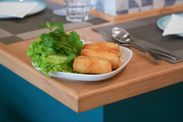 A white plate of crispy deep fried spring rolls wrapped stuffed with rice vermicelli and assorted veggies in a plate with a dipping sauce with green vegetables. Spoon and fork on table