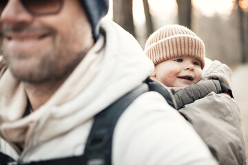 Sporty father carrying his infant son wearing winter jumpsuit and cap in backpack carrier hiking in...