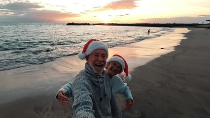 Two old happy seniors wearing christmas hats at the beach taking a selfie of them smiling and having fun with the sunset at the background at evening. Cute couple of old persons looking at the camera - Powered by Adobe