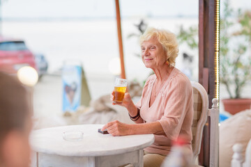 Senior woman sitting at a table in a summer cafe and drinking beer from a tall glass