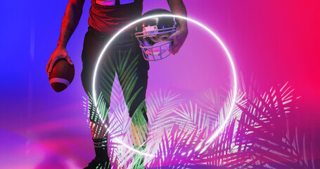 Low section of american football player holding ball and helmet by illuminated circle and plants