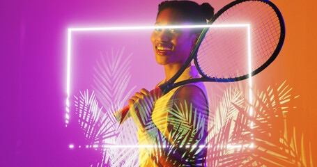Biracial smiling female player with tennis racket standing by illuminated rectangle and plants