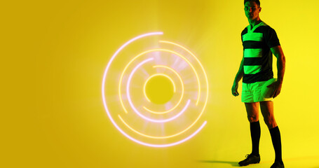 Full length of caucasian rugby player with ball standing by illuminated circular pattern, copy space