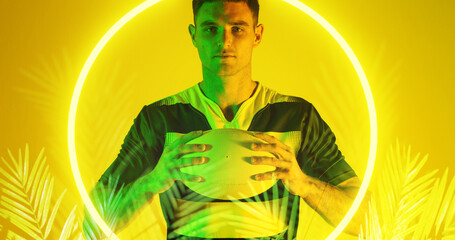 Caucasian male player holding rugby ball over illuminated circle and plants on yellow background