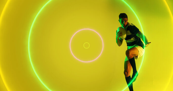 Illuminated colorful circles over caucasian male player with ball playing rugby on yellow background - Powered by Adobe