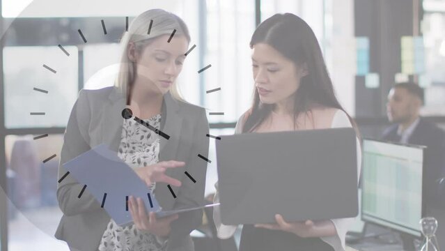 Animation of clock over two diverse businesswomen using laptop talking in office