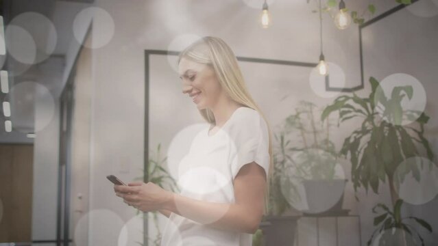 Animation of bokeh light spots over happy caucasian woman using smartphone in office