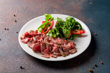 Sliced roast beef with green salad on white plate on dark stone table