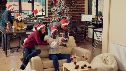 Cheerful colleagues playing video games at christmas party in office, feeling happy and enjoying winter holiday celebration. Using controllers on console to play gaming competition at festivity.