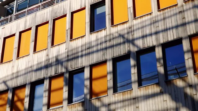 Orange window shades and timber cladding on the exterior wall, with shadows cast by adjacent office building. Using wood as material in modern architecture. Blue sky reflection in the glass.