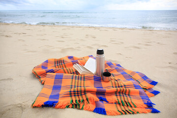 Metallic thermos with hot drink, open book and plaid on sandy beach near sea