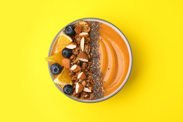 Bowl of delicious fruit smoothie with fresh orange slices, blueberries and granola on yellow background, top view