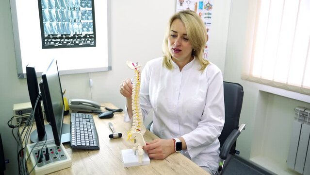 Neurologist explaining the functioning of a spine with the help of a plastic model. Smiling positive female medic in white robe working in the light cabinet.