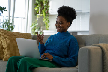 Happy sociable African American woman making video call in laptop sits on sofa. Positive charismatic girl with portable computer on knees leads online webinar and communicates with blog subscribers
