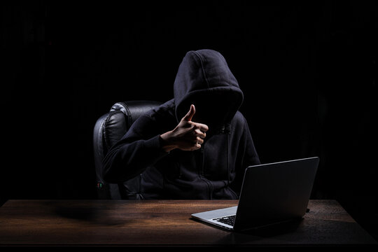 A hacker, dressed in a black hood, is raising a thumbs up when completing tasks to hack into a darkened room.