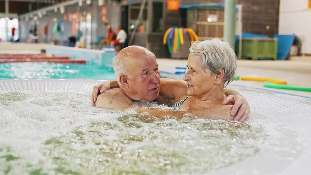 Senior caucasian married couple with swimming pool equipment - hair cap and glasses - hugging and talking while sitting in a hot-water . High quality 4k footage