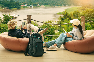 Male tourist wear hat with face mask and his wife use mobile phone and sit to relax on large cushions on balcony with mountains and river in background. Traveller in bag chair enjoy outdoor nature.