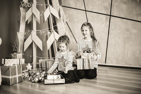 Little Sisters Are Looking At Christmas Presents Sitting In The Modern Living Room. Black And White Photo