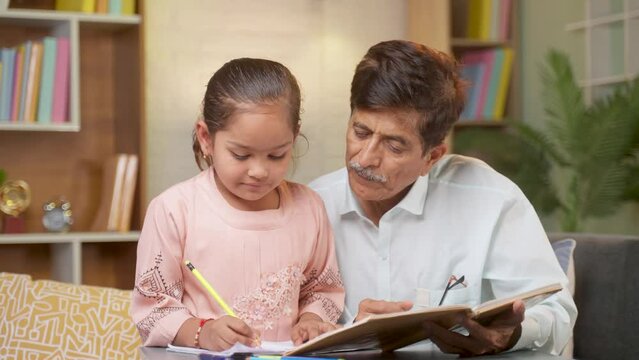 Grandfather helping his granddaughter to study at home by teaching from textbook - concept of family support, development and education