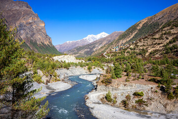 View of the valley in the mountains of Annapurna with a blue sky. A blue river flows through the valley and we see green trees on the slopes.