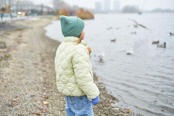 Girl standing at the coast and watching swimming birds on river. autumn misty grey river and cloudy sky landscape with wild ducks floating.