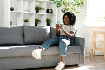 Fototapeta premium Smiling african woman sitting on couch at home and using smartphone