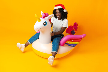 African woman sitting on rainbow unicorn float wearing santa claus hat in studio