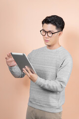 Handsome young asian man holding digital tablet and looking at it with smile while standing against beige background