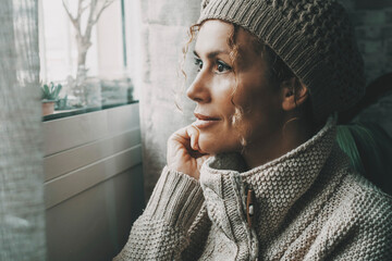 Fototapeta na wymiar Side portrait of serene female looking outside the window at home in winter season Cold house temperature. One woman wearing knit hat inside apartment. Relax and feeling expression lady leisure